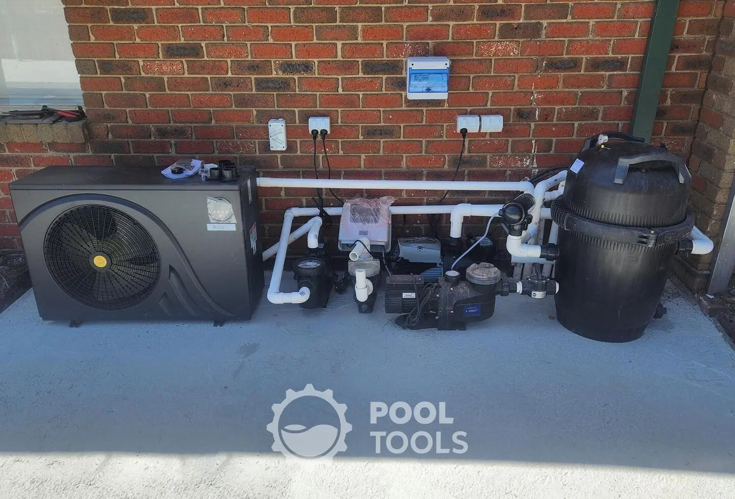 Pool equipment including a heat pump, filter, and control panel on a concrete surface with a brick wall background.
