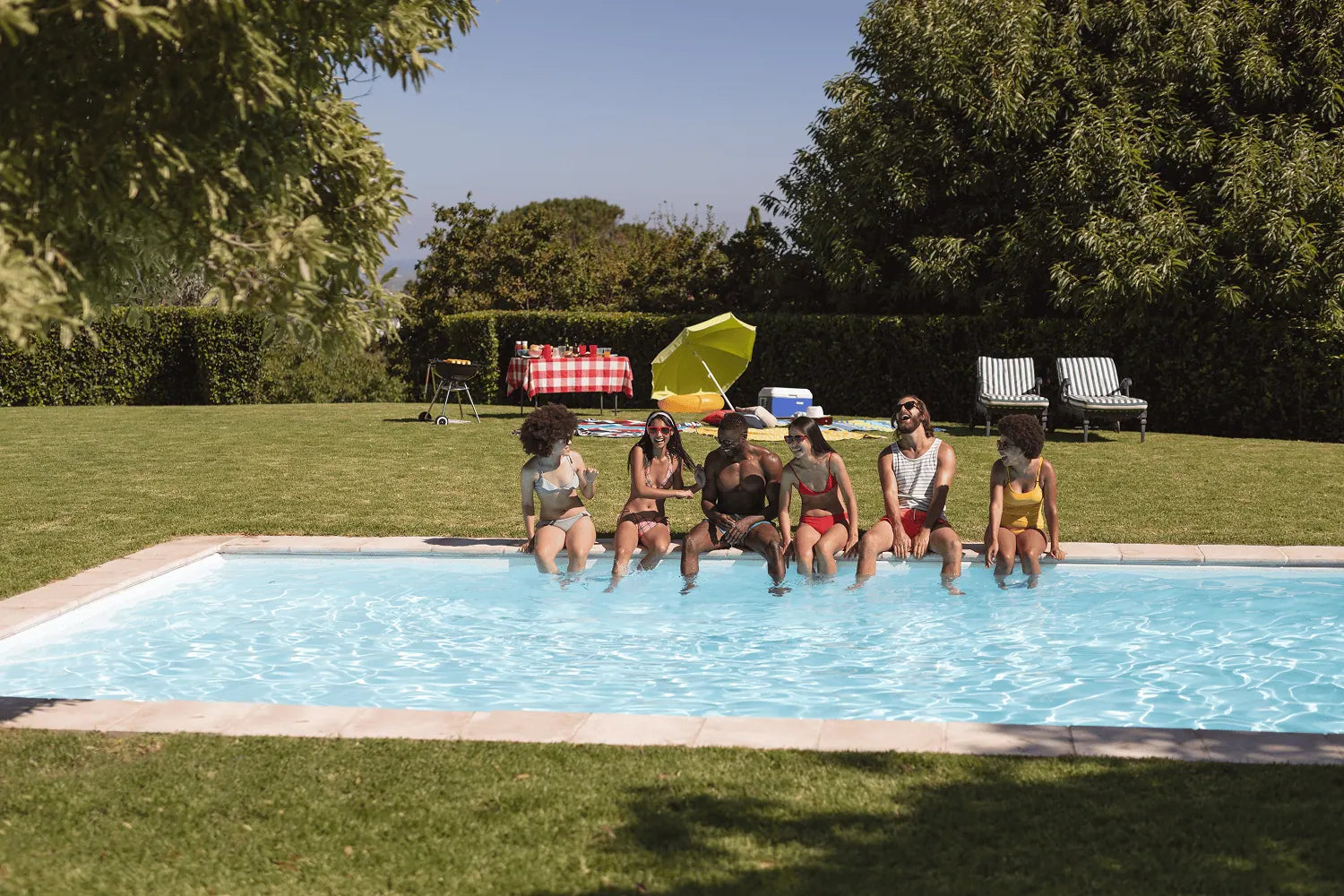 Group of people sitting by a pool with a colorful umbrella and chairs in the background.